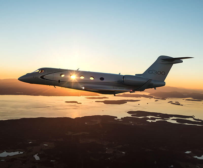 A Gulfstream G300 backlit by a rising sun flies above a distant archipelago.