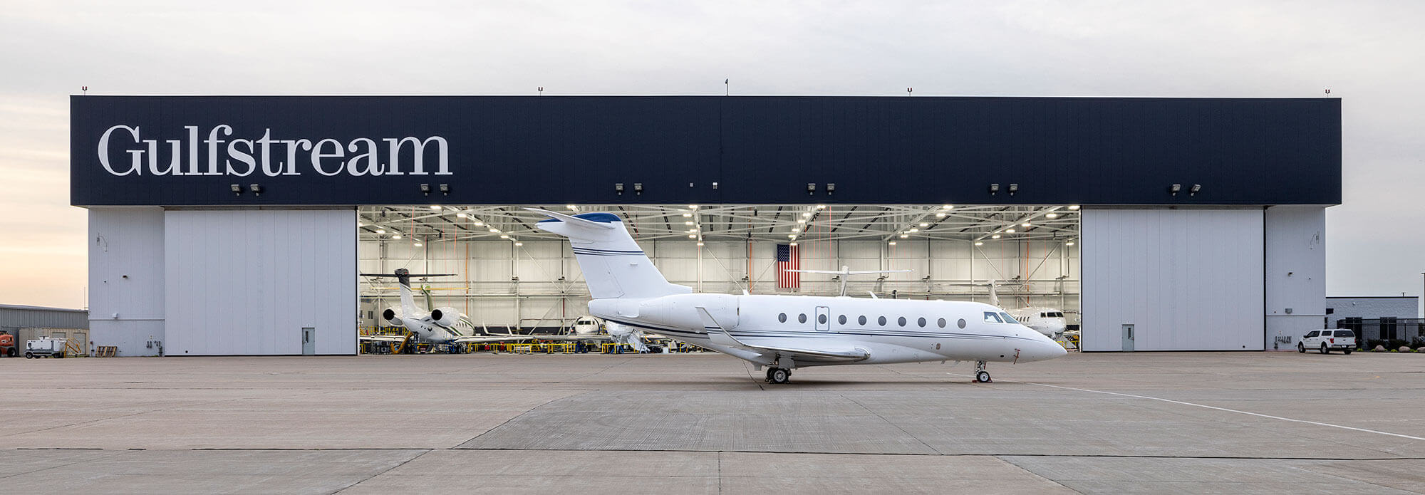 A Gulfstream G280 parked on the tarmac at dusk in front an open Customer Support service hangar door.