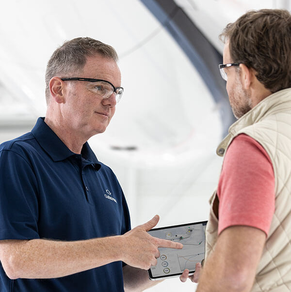 An employee wearing safety goggles in a service hanger presents information on an tablet screen to a customer.