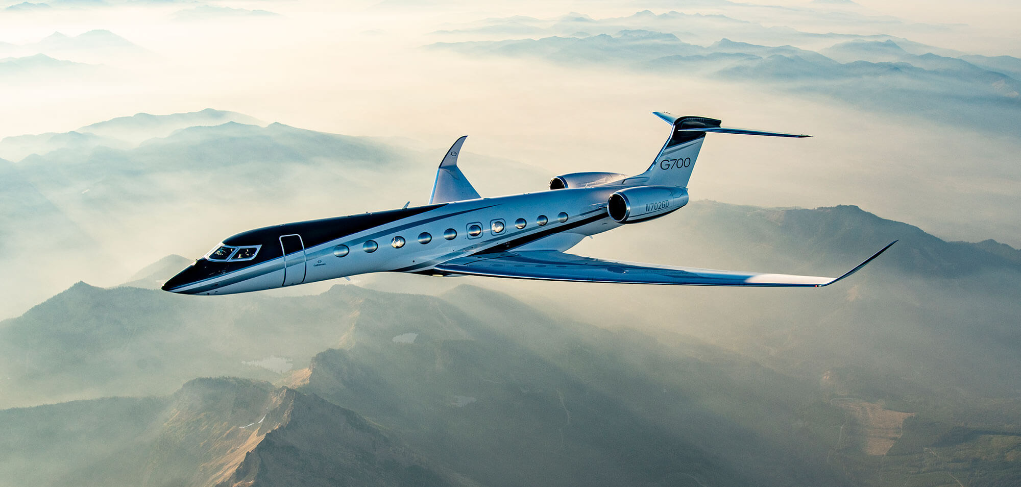 A Gulfstream G700 seen from above flies above snow-capped mountains far below.